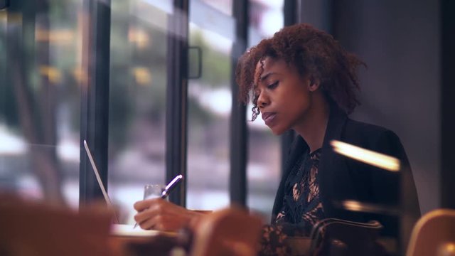 Side view of one pretty young African businesswoman busy working with laptop in the office by the window, a young female freelancer working in the cafe surfing the internet and writing notes