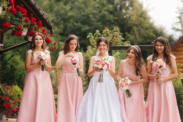Fototapeta premium Happy bride with bridesmaid hold bouquets and have fun outside. Beautiful bridesmaid in same dresses stand by the charming bride in long wedding dress