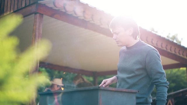 young man, standing near summer house, prepares the coals in the grill, fanning them.