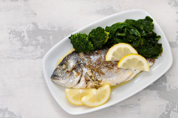 baked fish with lemon and vegetables in white dish on ceramic background
