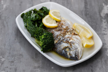 baked fish with lemon and vegetables in white dish on ceramic background