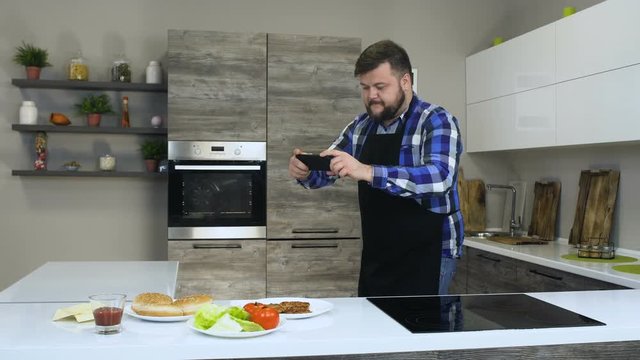 Thick bearded guy takes a pictures of Ingredients for homemade hamburger using a phone in a kitchen, before cooking a homemade burger