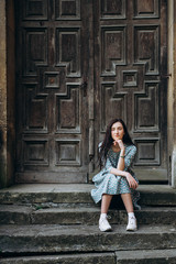 Fototapeta na wymiar Attractive young woman sitting near old doors in cute summer dress with flower print