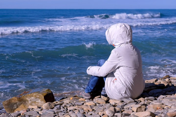 Woman in a white down jacket and hood sits on the shore and looks at the stormy sea waves beating against the stone shore.