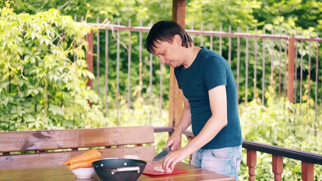 Young guy is cooking pilaf. He is in a summer house, cuts meat, on a red cutting board. He throws it into the cauldron. Preparing for a picnic.