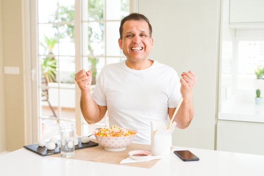 Middle age man eating asian food with chopsticks at home celebrating surprised and amazed for success with arms raised and open eyes. Winner concept.