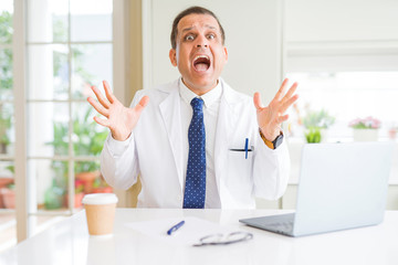 Middle age doctor man wearing white medical coat working with laptop at the clinic celebrating crazy and amazed for success with arms raised and open eyes screaming excited. Winner concept