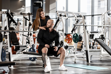 Fototapeta na wymiar Young slender pretty girl stands behind the athletic man sitting on the spot bench and holds her hands on his shoulders in the modern gym