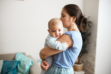 Fototapeta na wymiar Young mother dressed in light blue t-shirt and skirt is holding her tiny son on her arms in the room at home