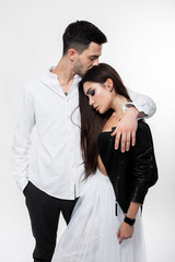 Beautiful brunette girl in a white tulle skirt in a black bra and black leather jacket with guy in trousers and white t-shirt are hugging on the white background in the studio