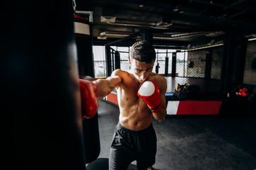 Sportsman in red boxing gloves with a naked torso hits punching bag in the gym on the background of  boxing ring