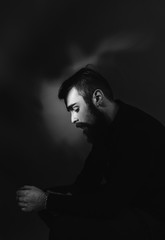 Black and white photo of a man with a beard and stylish hairdo dressed in the black shirt sitting on the stool in the fog background