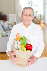 Handsome senior man holding paper bag full of fresh groceries and smiling at home