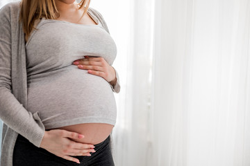 Pregnant woman touching her belly standing by the window at home