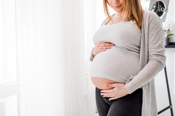 Pregnant woman touching her belly standing by the window at home