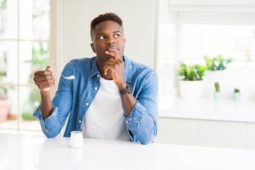 African american man eating healthy natural yogurt with a spoon serious face thinking about...