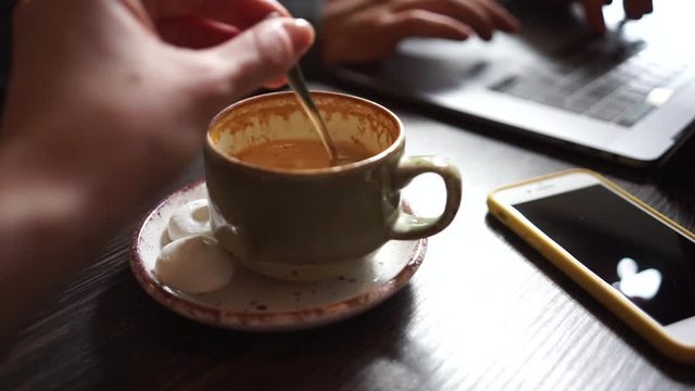 Close up cup of coffee on wooden table. Woman hand stir the sugar. Smartphone, keyboard and tapping fingers on the background