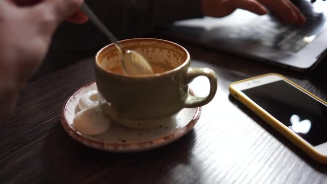 Close up cup of coffee on wooden table. Woman hand stir the sugar. Smartphone, keyboard and tapping fingers on the background