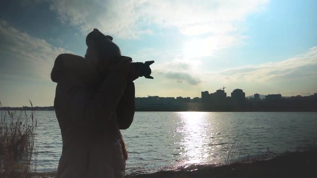 Young woman taking pictures of the sunset on the background of the sea, city and sky. Female silhouette takes pictures while holding a professional camera lens. Overall plan.