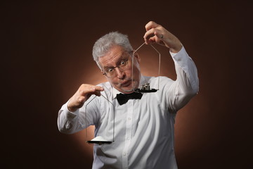 Elderly gray-haired man 50s, in white shirt, glasses and bow tie weighing something on scales with kettlebells