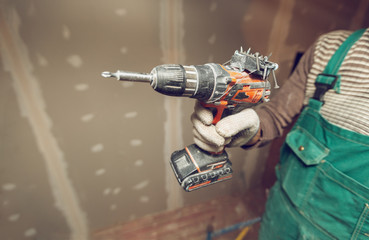 Worker with protective gloves is holdind a perforator with some drywall screws in apartment that is...