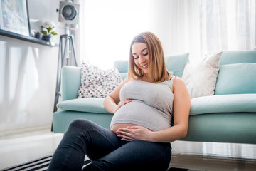 Beautiful smiling pregnant woman sitting on floor at home, touching and looking on belly
