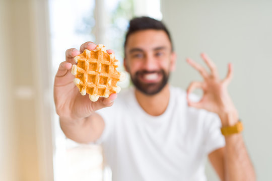 Handsome hispanic man eating sweet belgian waffle pastry doing ok sign with fingers, excellent symbol