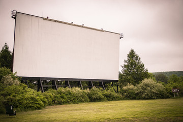 Drive-in theater movie screen stands in the distance; surrounded by grass and wooded overgrowth.