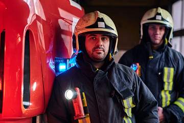 Two firemen wearing protective uniform standing next to a fire truck in a garage of a fire...