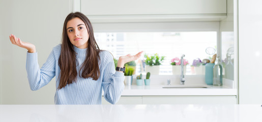 Wide angle picture of beautiful young woman sitting on white table at home clueless and confused expression with arms and hands raised. Doubt concept.