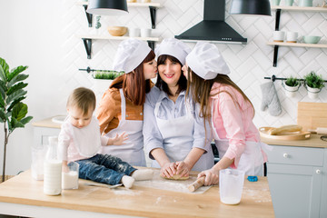 Young woman and her sister, middle aged woman and little cute daughter preparing the dough, bake cookies at Mothers Day. Daughters are kissing her mother
