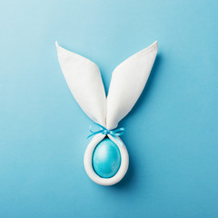 Easter bunny ears made of white napkin with egg on blue background. Minimal styled easter card concept.