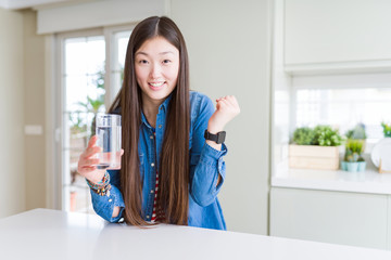 Beautiful Asian woman drinking a glass of fresh water screaming proud and celebrating victory and success very excited, cheering emotion