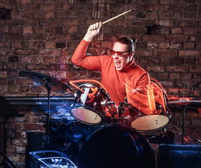 Fototapeta na wymiar Young Stylish musician in sunglasses emotionally playing drums against brick wall background, perform in a night club. Photo with lighting motion effect