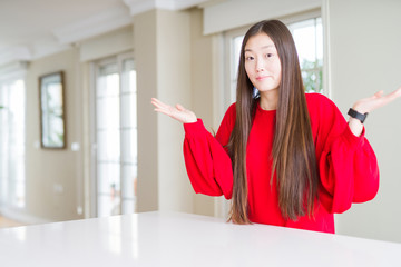 Beautiful Asian woman wearing red sweater on white table clueless and confused expression with arms...