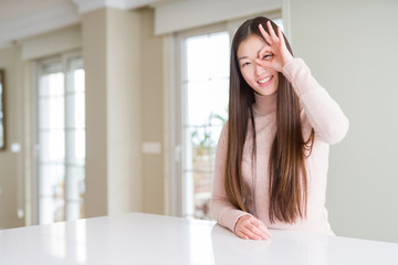 Beautiful Asian woman wearing casual sweater on white table doing ok gesture with hand smiling, eye looking through fingers with happy face.