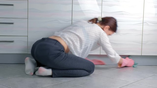 Woman in pink rubber gloves washes hard kitchen furniture with cloth, sitting on the floor. Back view.