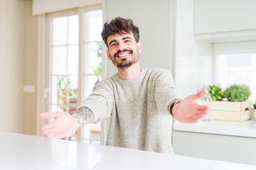 Fototapeta na wymiar Young man wearing casual sweater sitting on white table looking at the camera smiling with open arms for hug. Cheerful expression embracing happiness.