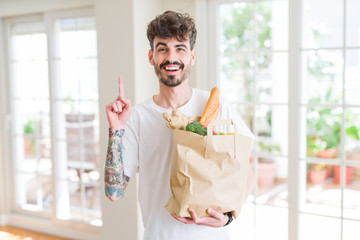 Young man holding paper bag of fresh groceries from the supermarket surprised with an idea or question pointing finger with happy face, number one