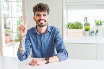 Young man wearing casual shirt sitting on white table showing and pointing up with fingers number two while smiling confident and happy.