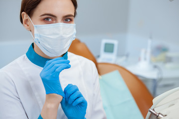 Close up of a female doctor wearing uniform and medical mask looking to the camera while putting on rubber gloves, copy space. Professional dentist at her office. Dentistry, dental care concept