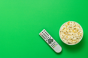A bowl of popcorn and a TV remote on a green background. The concept of watching TV, film, TV series, sports, shows. Flat lay, top view.