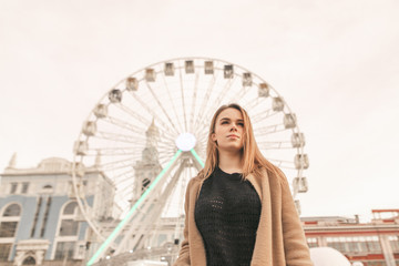 Serious attractive girl in spring clothes, wearing a beige coat, standing in front of the street and ferris wheel, looking forward. Street portrait of a beautiful young woman on a walk