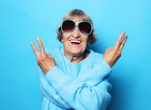 Funny old lady wearing blue sweater, hat and sunglasses showing victory sign. Isolated on blue background.