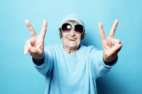 Funny Old Lady Wearing Blue Sweater, Hat And Sunglasses Showing Victory Sign. Isolated On Blue Background.