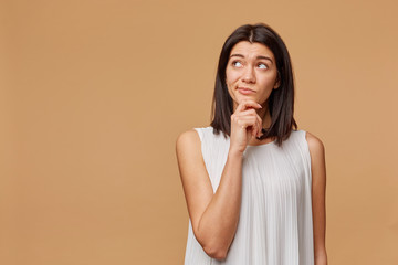 Unsatisfied thoughtful tanned woman standing hand holding chin, frowning, looking at the upper left corner at the blank copyspace, expressing suspiciousness and misunderstanding, over beige background
