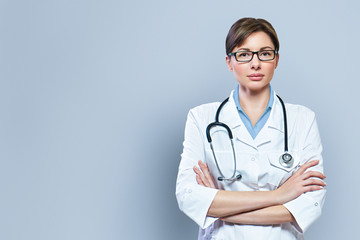 Portrait female doctor in white uniform standing with crossed hands on blue clinic background