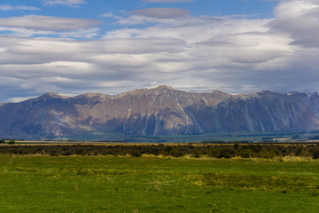 Mountains on the horizon covered with shade, dramatic sky during warm sunny day, trees and green grass in the foreground
