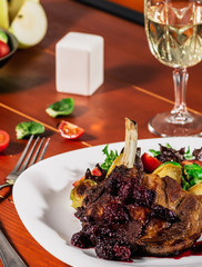 Roasted meat with roast apple, brussel sprouts, cherry tomatoes and salad leaves on a white plate and a glass of white wine, on wooden background