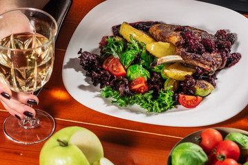 Roasted meat with brussel sprouts, cherry tomatoes, salad leaves and apple for garnish. girls hand with white wine